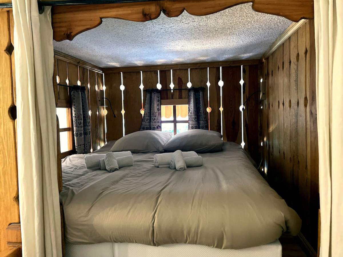 Intérieur de la cabane en bois avec lit double, rideaux et bois ajouré, couchage insolite de l’appartement à Serre Chevalier
