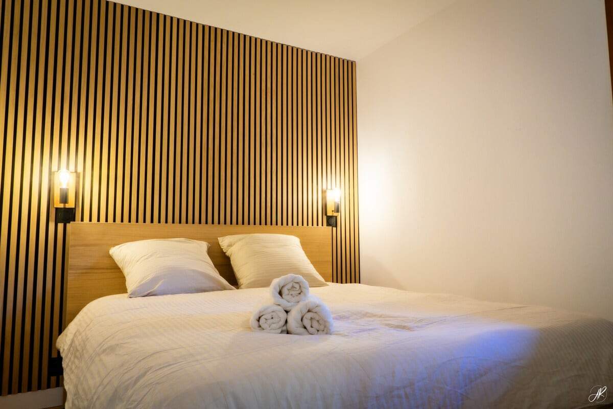 Chambre principale avec tête de lit en tasseaux de bois, appliques noires et serviettes roulées sur le lit