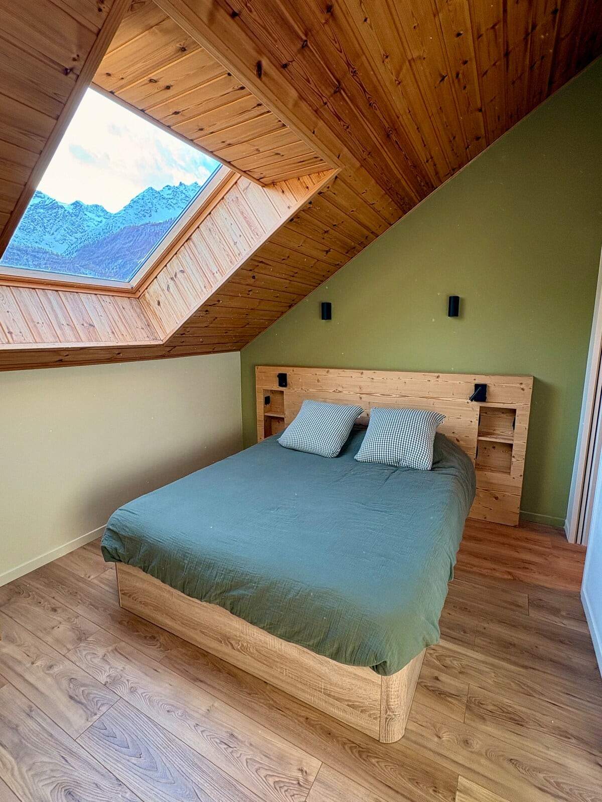 Chambre sous pente avec lit double, mur vert et velux ouvrant sur les sommets enneigés