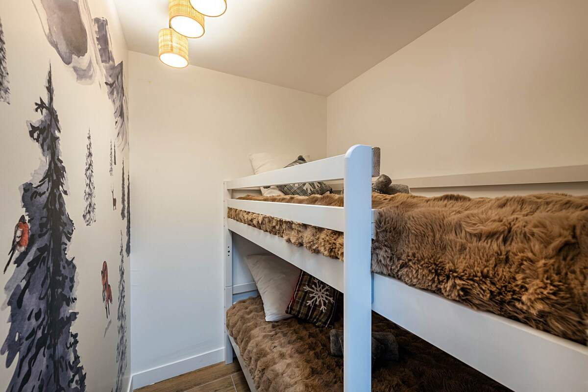 Coin nuit enfants avec lits superposés blancs, plaids en fourrure et fresque murale de sapins et d'animaux de montagne