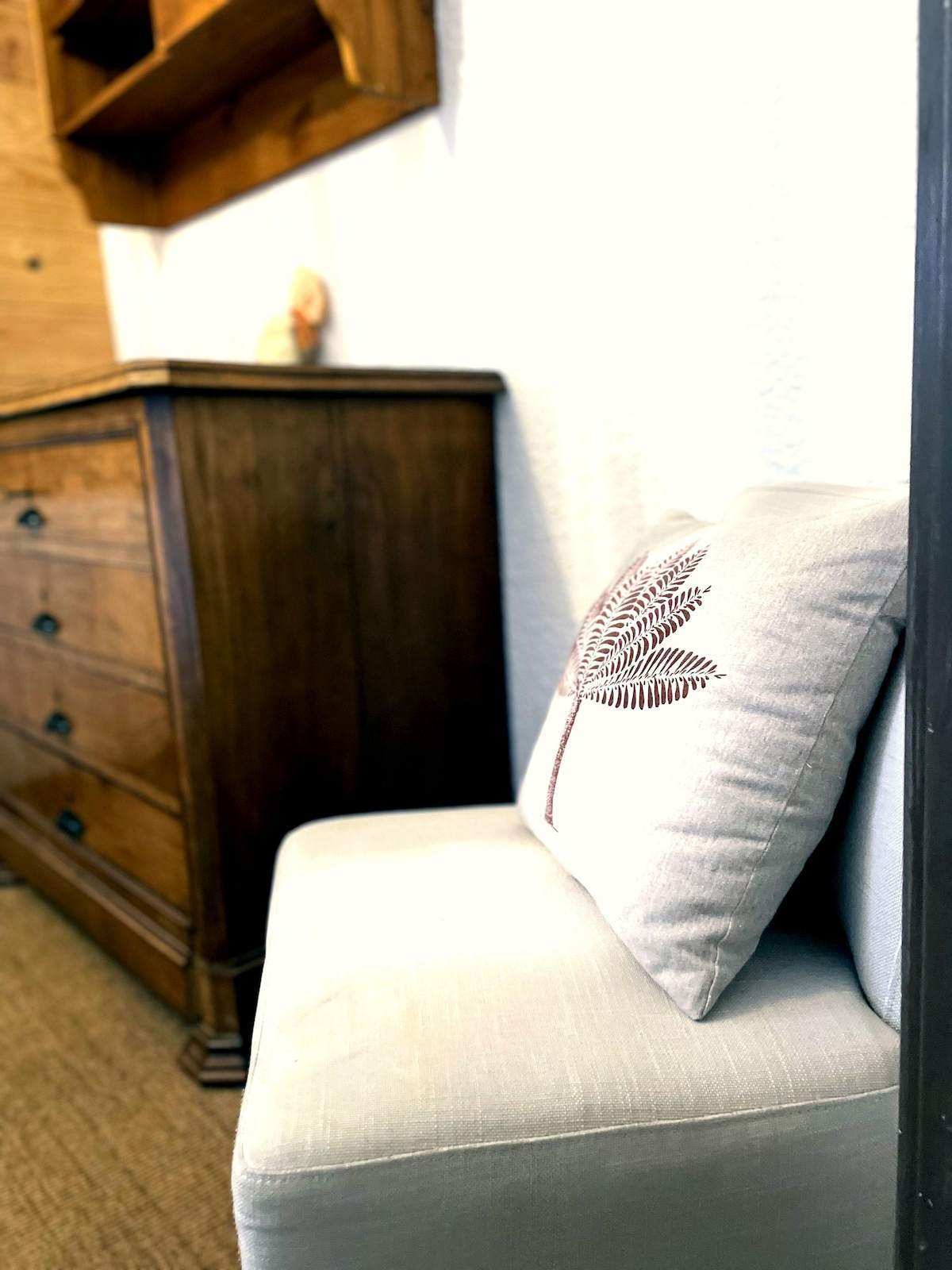 Détail de décoration : coussin brodé fougère sur une banquette en lin, à côté d’une commode ancienne