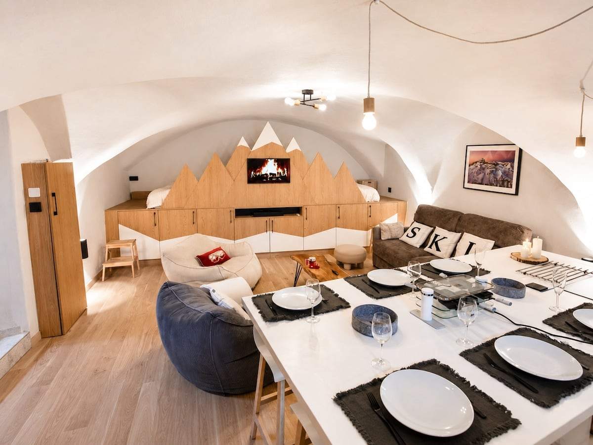 Pièce de vie voûtée du loft Le 1350 : îlot dressé pour six, fresque de montagnes en bois intégrant les couchages et la télévision, canapé et poufs