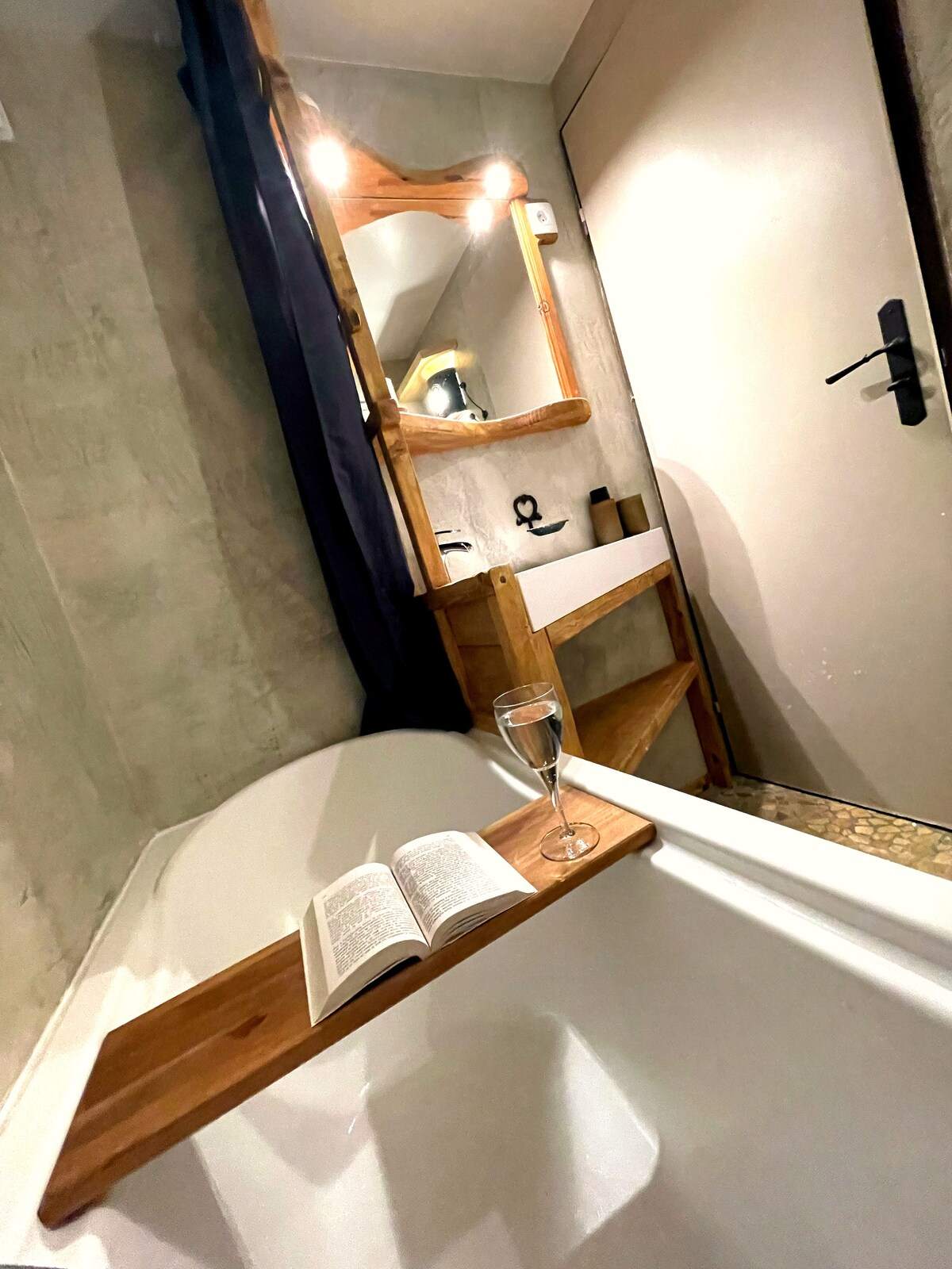 Salle de bain en béton ciré avec baignoire, plateau de bain en bois, livre et coupe de champagne, appartement à Serre Chevalier