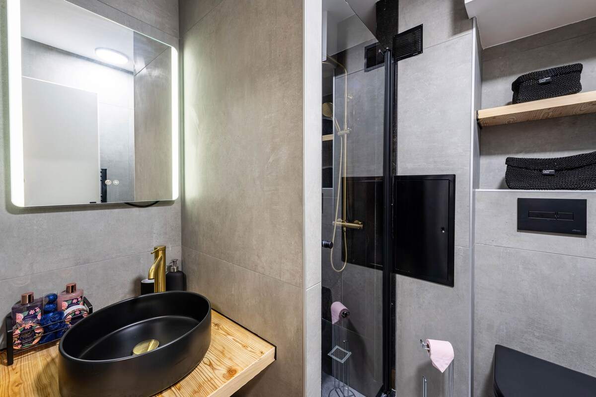 Salle de bain en béton ciré avec vasque noire, robinetterie dorée, miroir rétroéclairé et douche à l’italienne