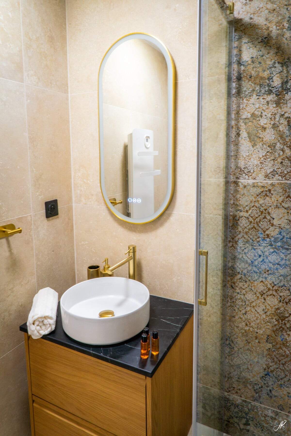 Salle de bain en travertin avec vasque ronde, robinetterie et miroir dorés, douche à l’italienne