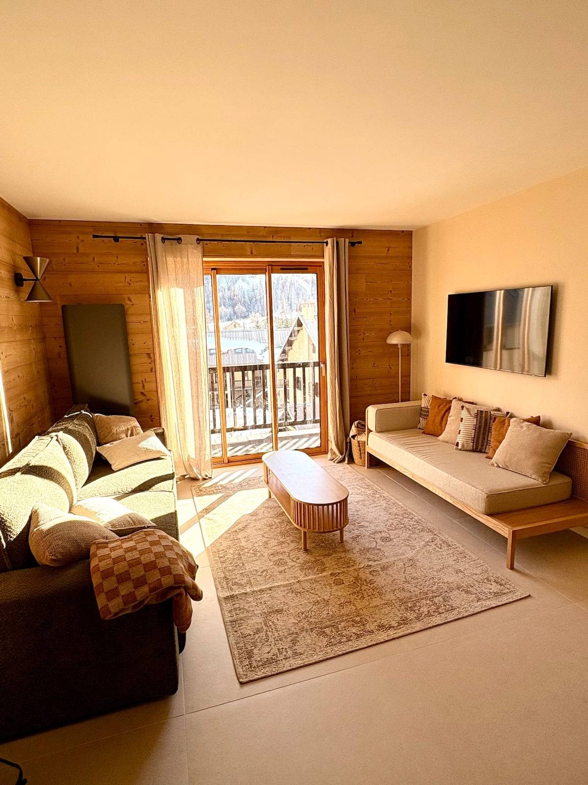 Salon de La Parenthèse Alpine baigné de soleil : lambris de mélèze, canapé, banquette en bois et grande baie vitrée ouvrant sur le balcon