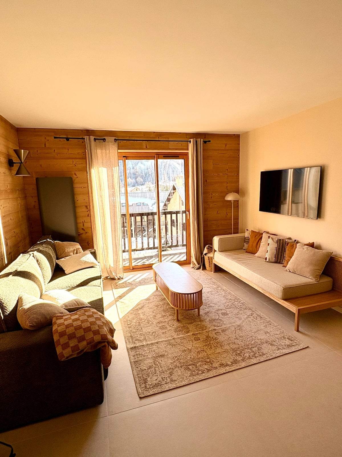 Salon de La Parenthèse Alpine baigné de soleil : lambris de mélèze, canapé, banquette en bois et grande baie vitrée ouvrant sur le balcon