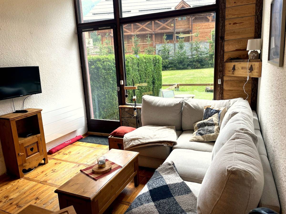 Salon de l'appartement à Serre Chevalier avec canapé d'angle en lin, grande baie vitrée ouvrant sur le jardin privatif et les chalets