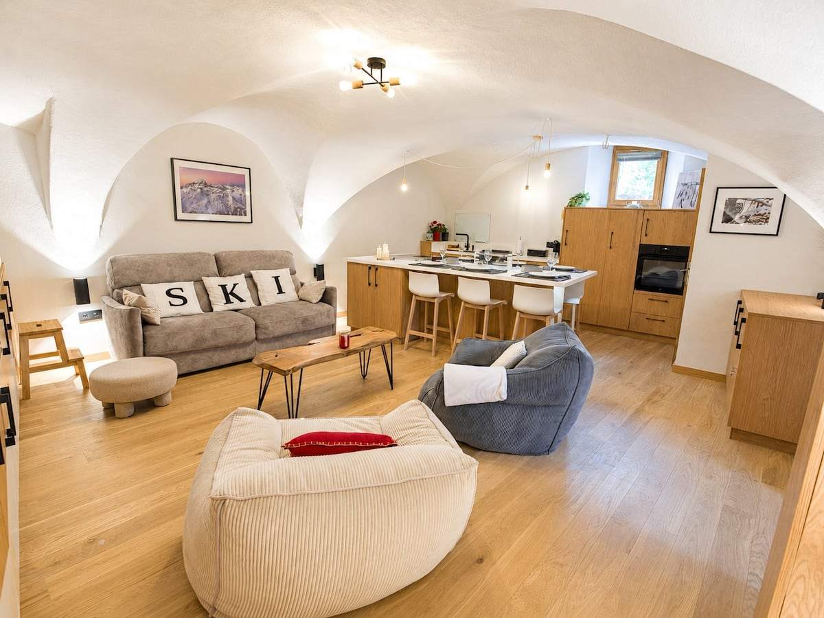 Salon du loft avec canapé, coussins brodés SKI, poufs en velours côtelé et îlot de cuisine en arrière-plan