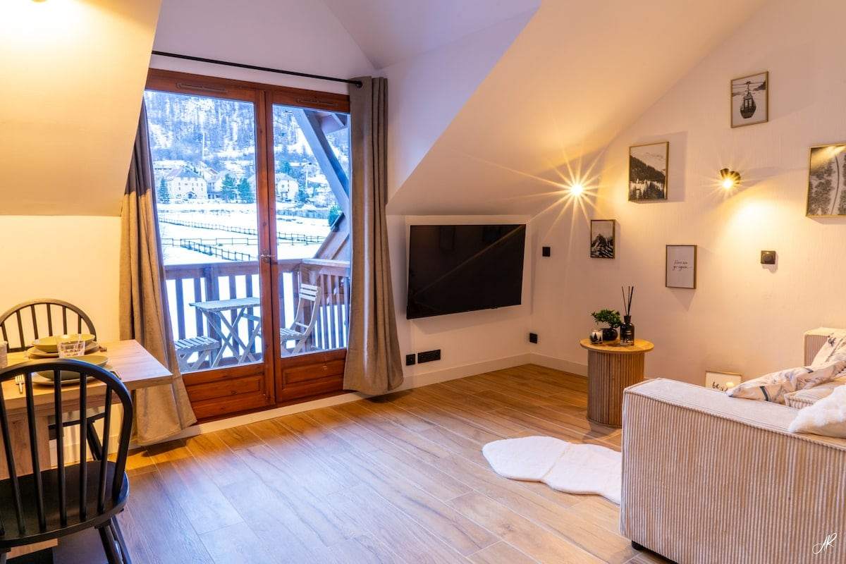 Séjour du Flocon sous pente, canapé clair et grande porte-fenêtre ouvrant sur le balcon et le village enneigé