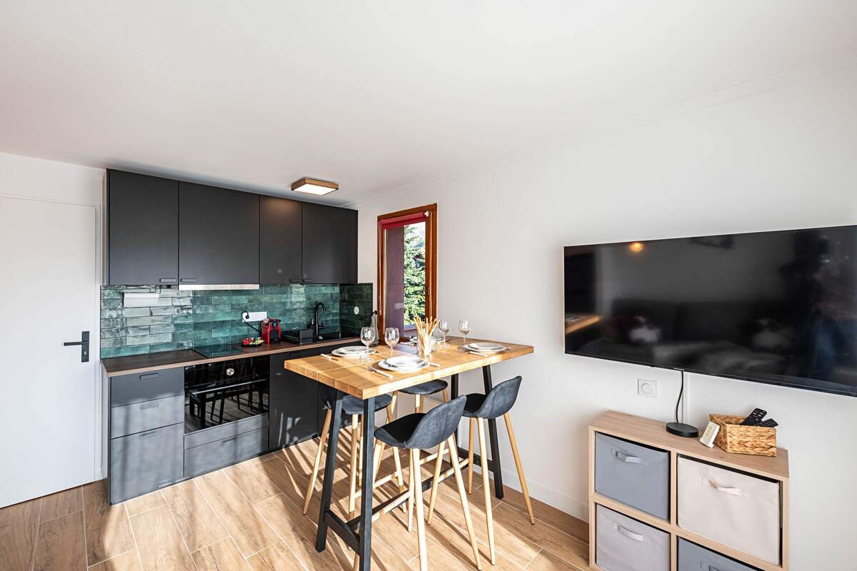Séjour lumineux avec cuisine équipée noire, table haute et tabourets, grand téléviseur mural et rangements