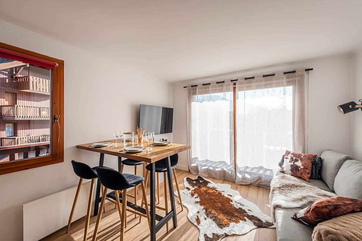 Table haute dressée face au canapé, tapis en peau de vache et baie vitrée voilée de l'appartement au pied des pistes