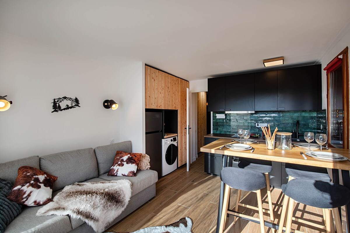 Séjour de l'appartement au pied des pistes à Serre Chevalier : canapé convertible gris et plaids en fourrure, cuisine noire à crédence verte et table haute en bois