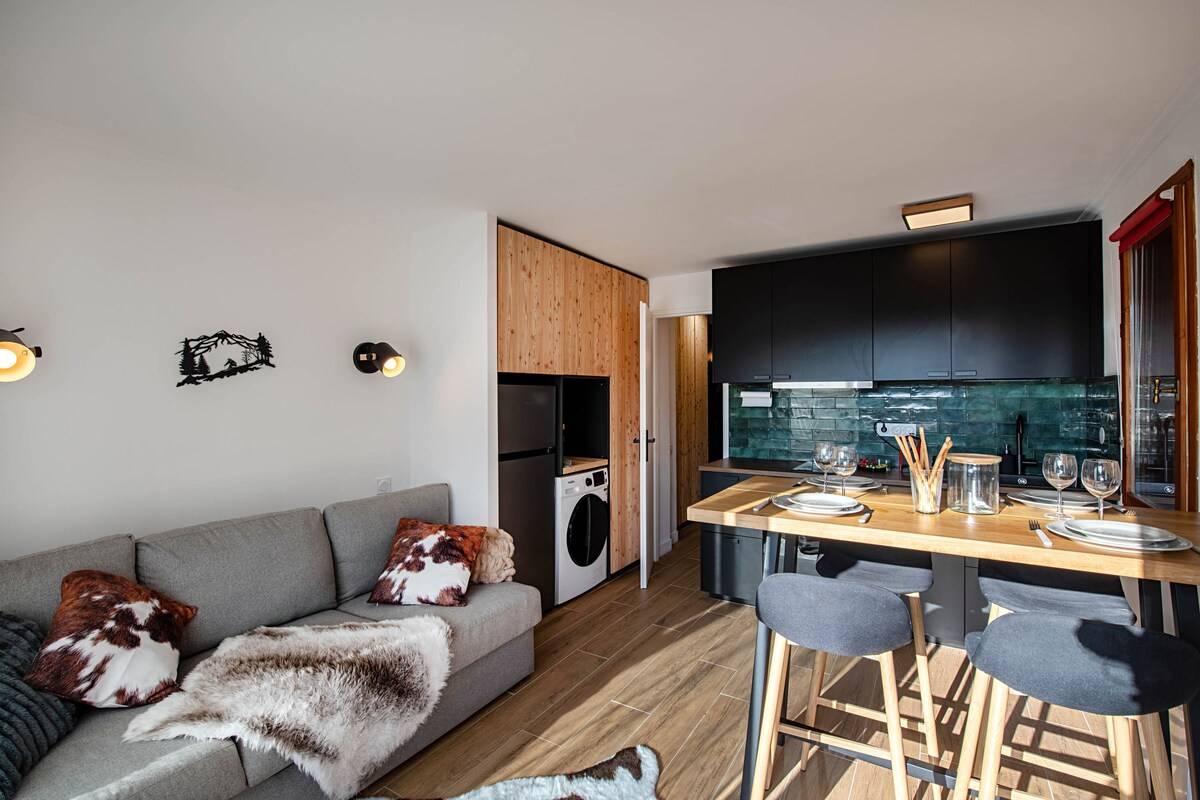 Séjour de l'appartement au pied des pistes à Serre Chevalier : canapé convertible gris et plaids en fourrure, cuisine noire à crédence verte et table haute en bois