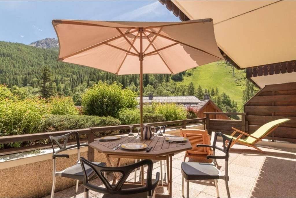 Terrasse plein sud de l'appartement de Chantemerle, table dressée sous un parasol, vue sur les pistes de Serre Chevalier en été